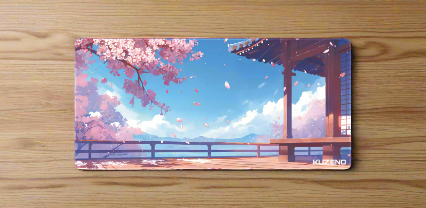 Sakura Skies Kuzeno Gaming Mousepad displayed on a wooden desk, featuring blooming cherry blossoms, a traditional pagoda, and serene mountains beneath a clear sky.